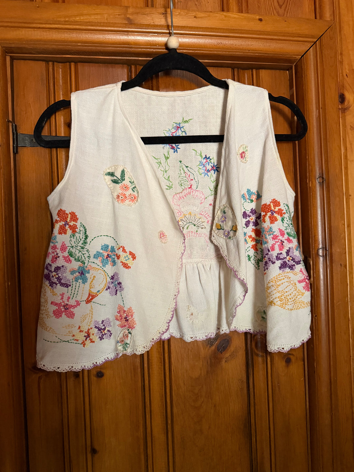 An off-white linen vest with colorful floral embroidery, hanging on a black hanger against a wooden door.
