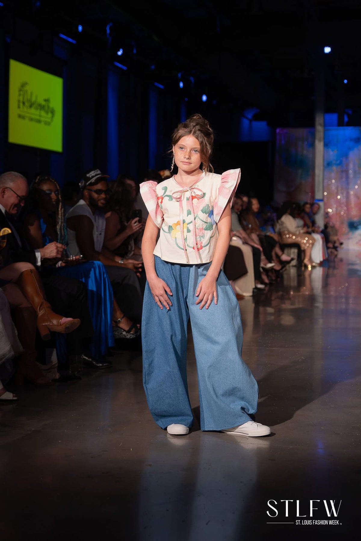A fashion runway photo shows a young girl walking down the catwalk wearing a white ruffled top with colorful floral embroidery, wide-leg blue jeans, and white sneakers. The runway is lined with seated spectators under stage lighting.