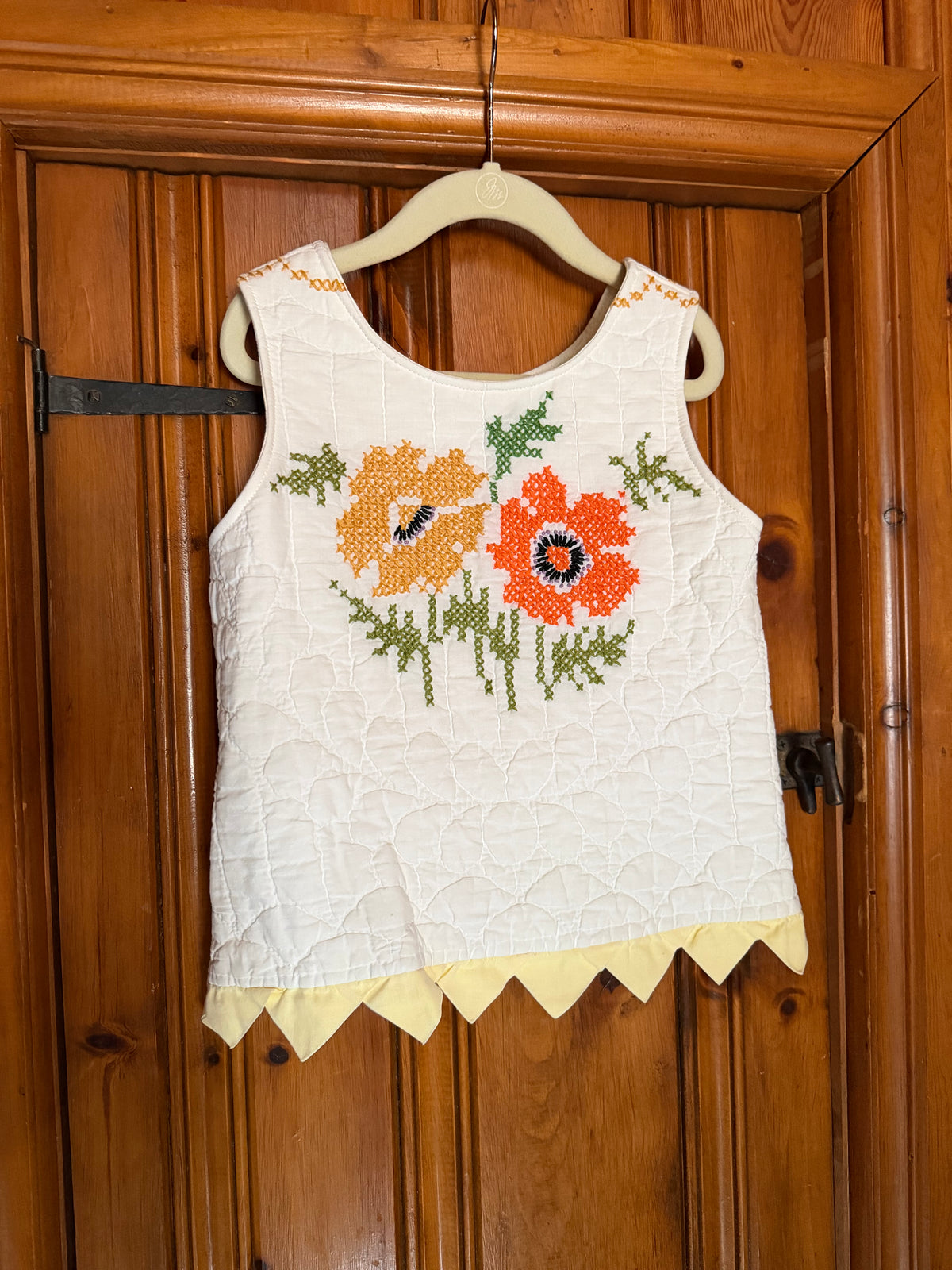 A white sleeveless quilted top with embroidered orange and yellow flowers and green leaves, hanging on a wooden hanger against a wooden door.