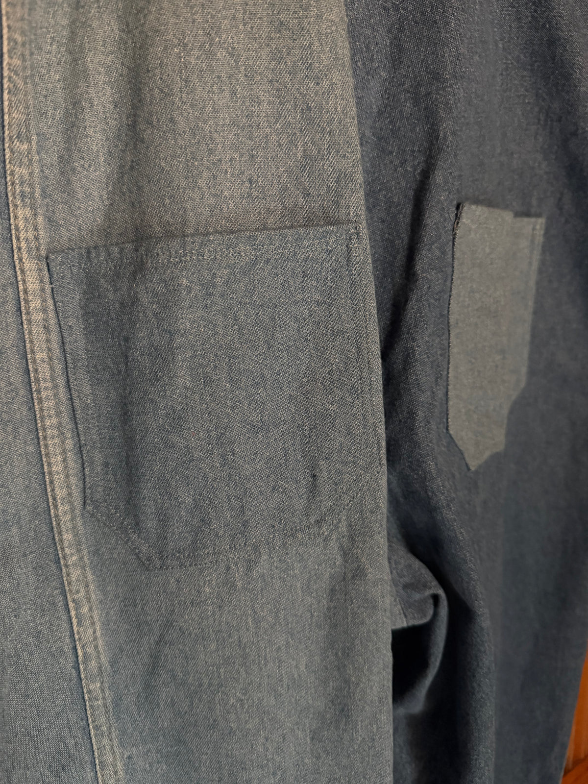Close-up of a dark gray patchwork shirt or overshirt featuring a large front patch pocket and a smaller side pocket, made from contrasting gray and navy-toned fabrics.