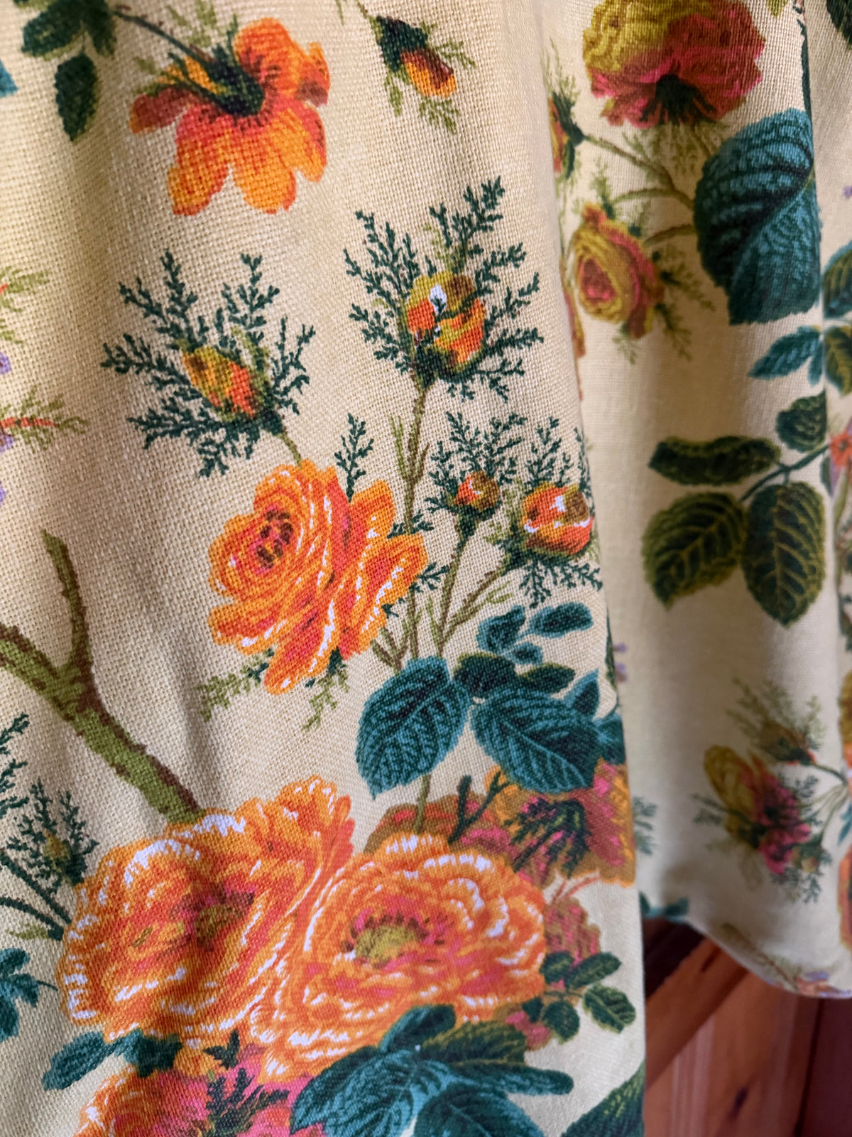 Close-up of a floral textile with large orange and peach roses, green leaves, and dark stems on a warm cream background.