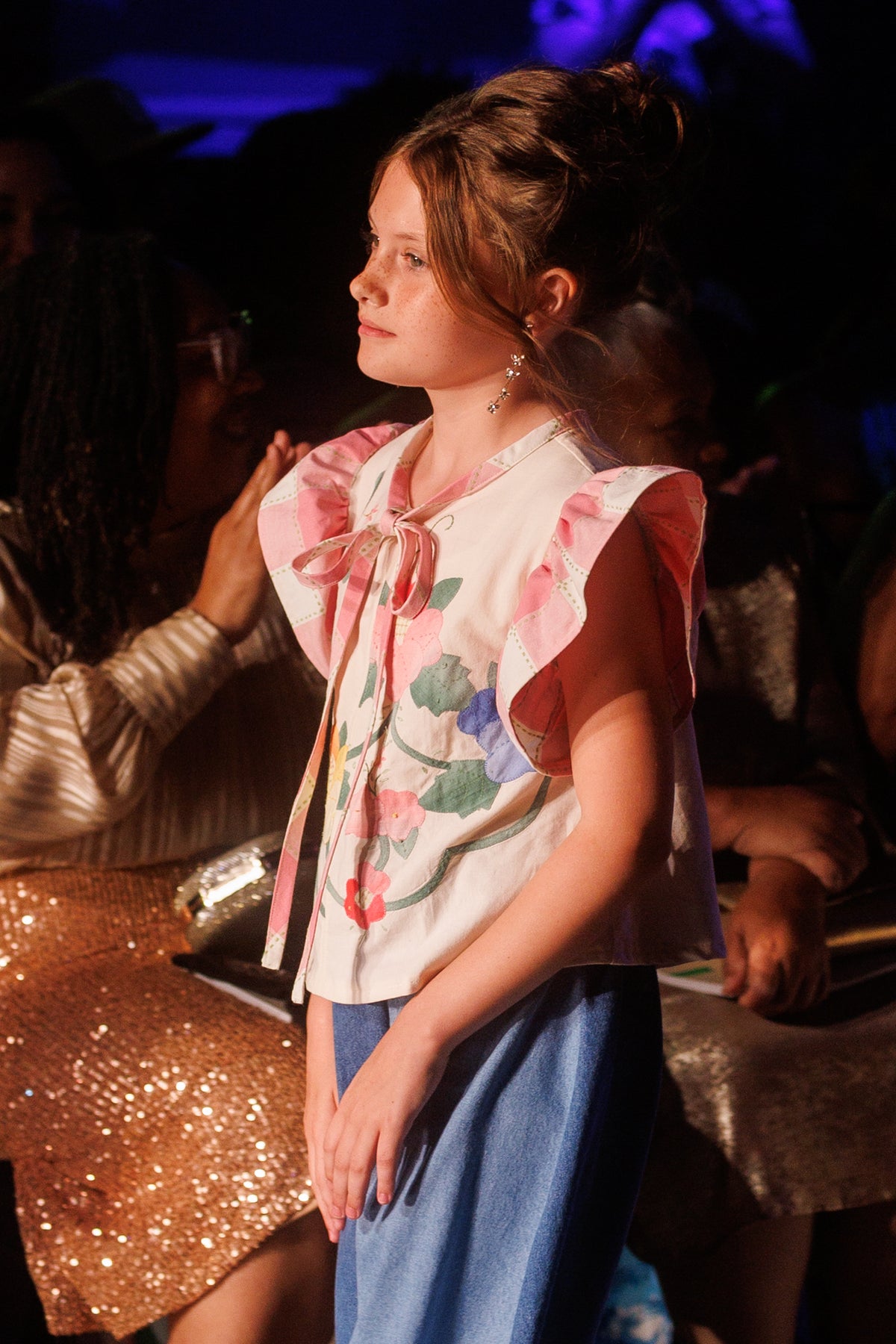 A young girl stands on a runway wearing a white ruffled blouse with pink and green floral embroidery and a blue skirt. The photo is taken on a fashion show with an audience in the background.