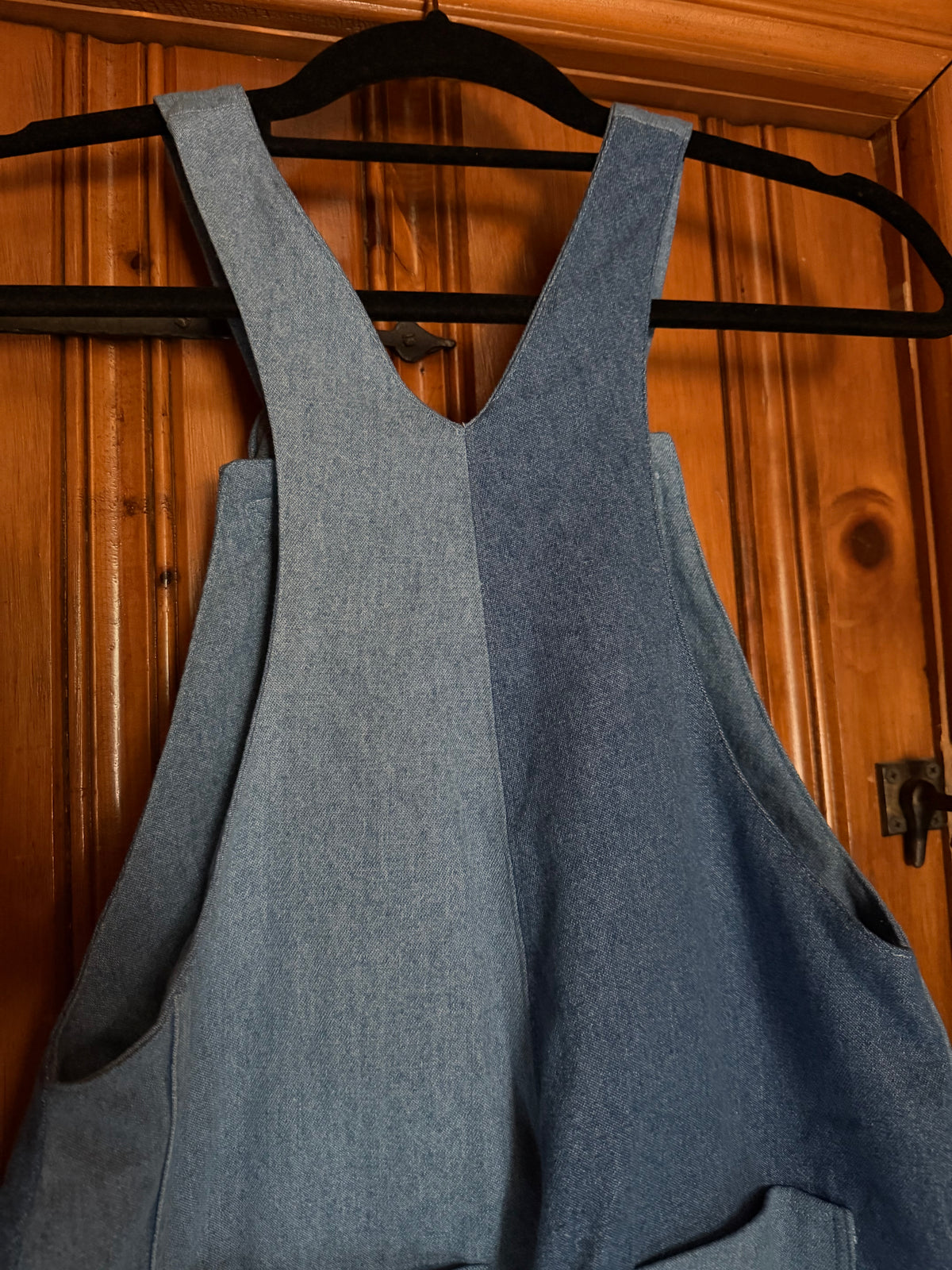 A pair of blue denim overalls  hanging on a black hanger against a warm wooden door background. Back view. 