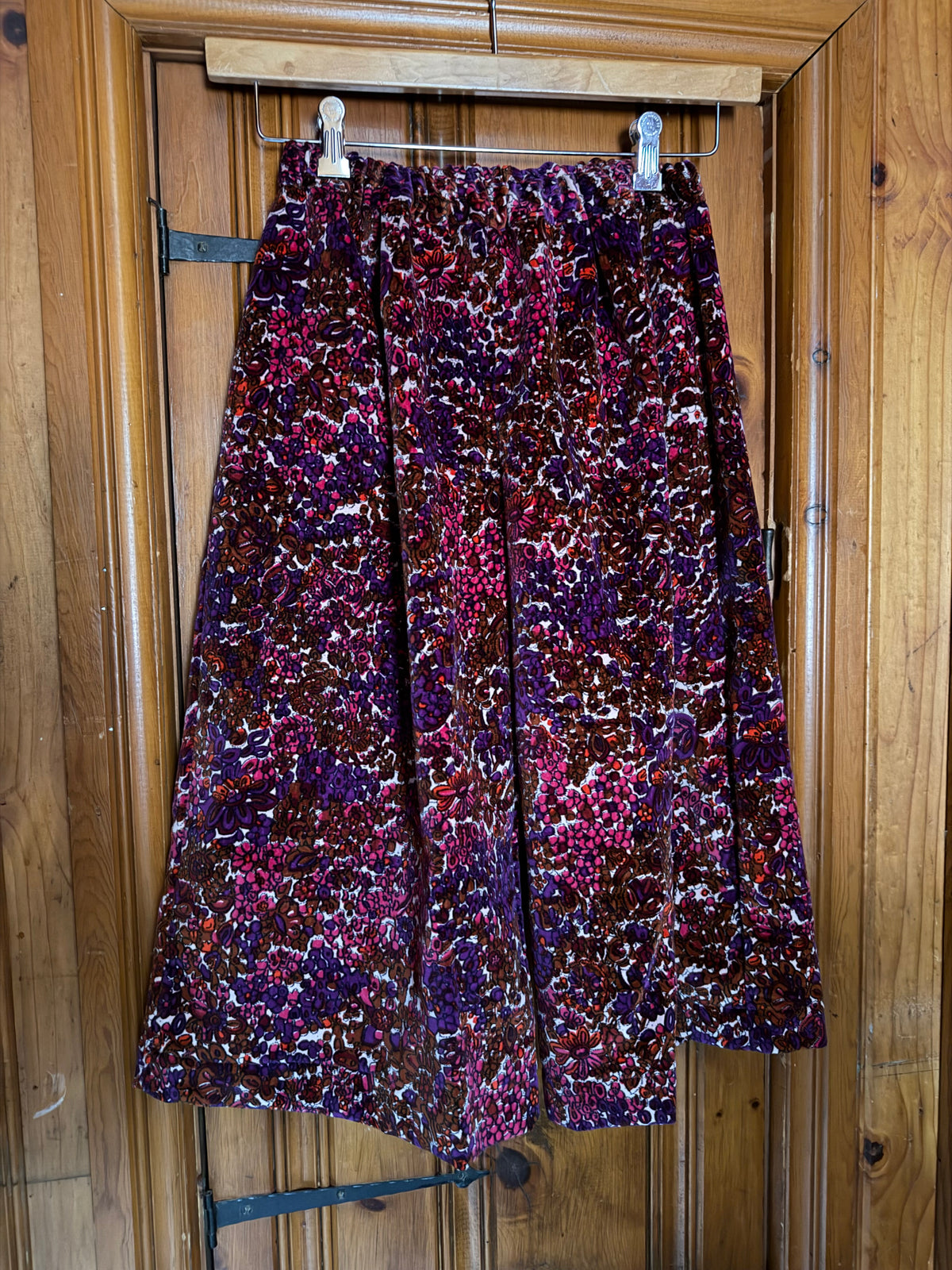 A pair of gaucho with a dense abstract floral pattern in purple, magenta, red, and brown, hanging on a wooden hanger against a wooden door.
