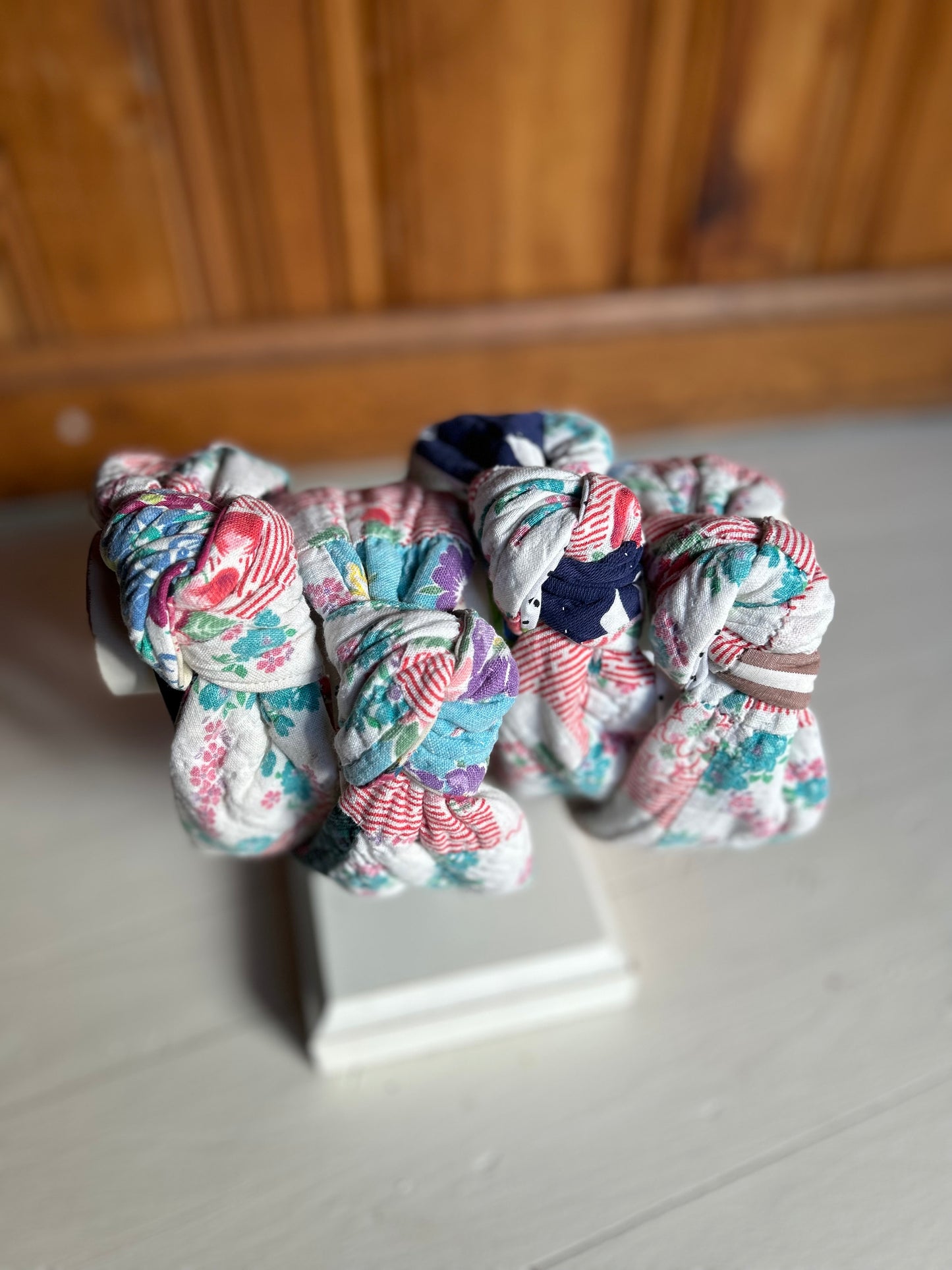 Image is of four knot headbands constructed with a vintage multi color quilt. Primary colors consist of a base color of white with red, greens and blues. The headbands are hanging on a white wooden necklace holder.