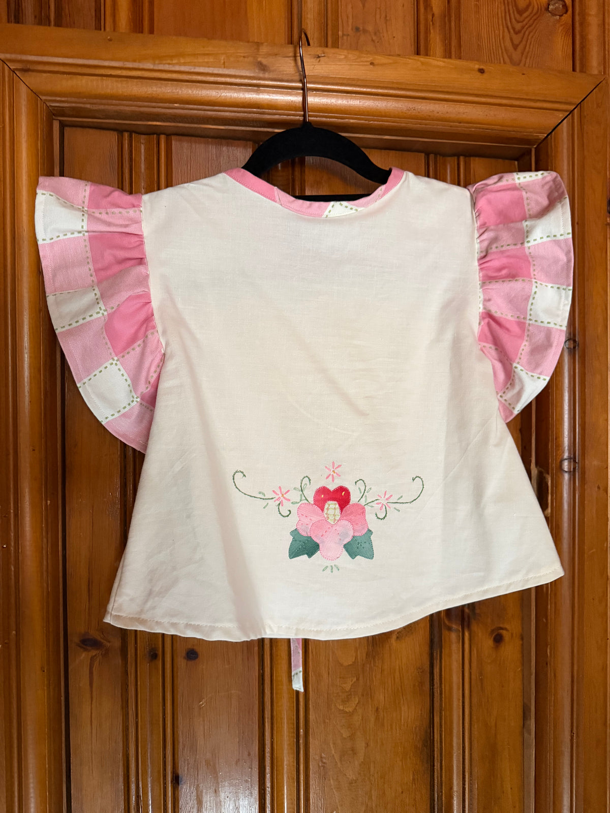A cream-colored children's top with pink ruffled flutter sleeves and a pink and green floral embroidery on the back, hanging on a black hanger against a wooden door.