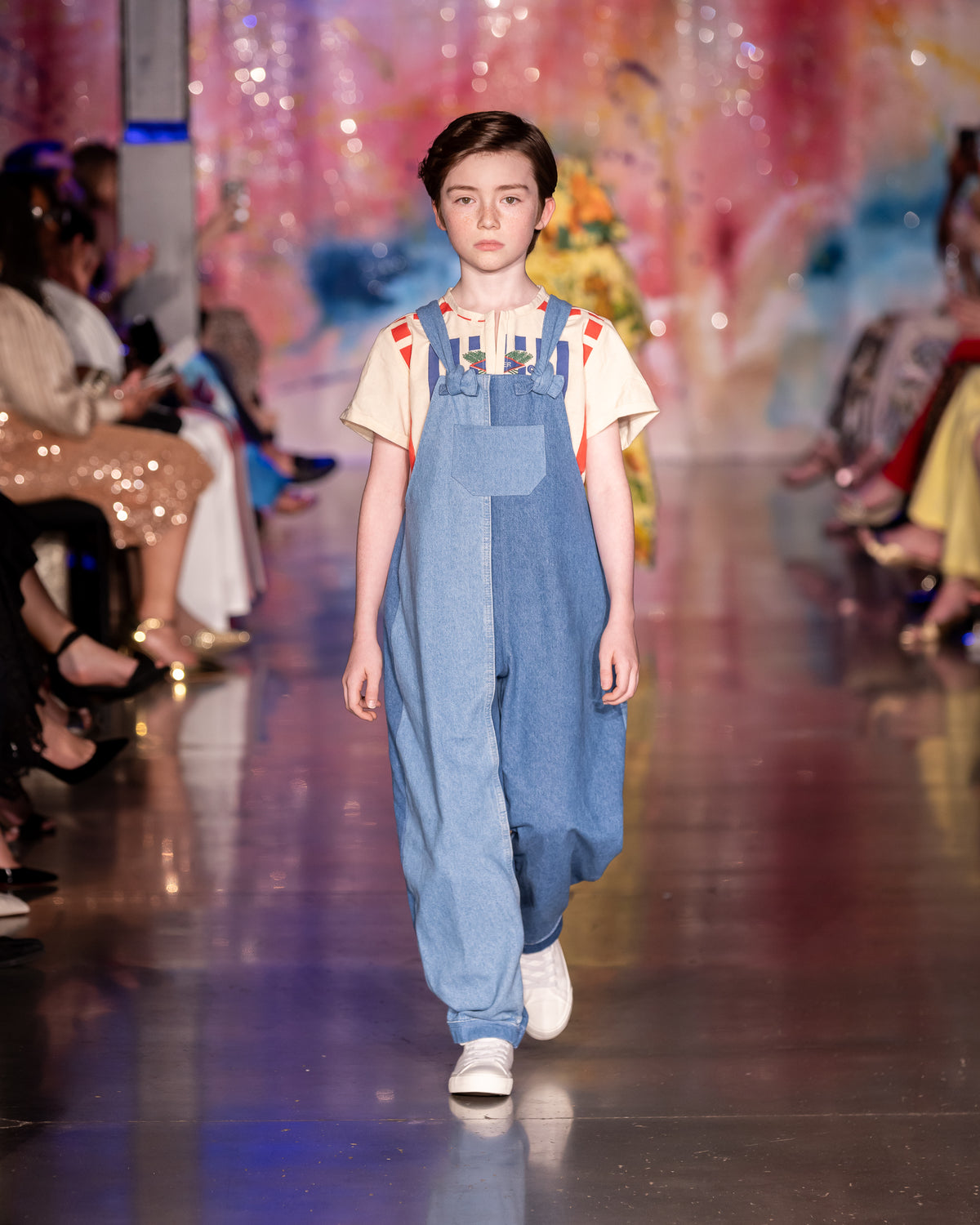 A child model walks down a fashion runway wearing blue denim overall-style pants over a cream short-sleeve top with small red accents, with seated audience members visible on both sides of the catwalk.