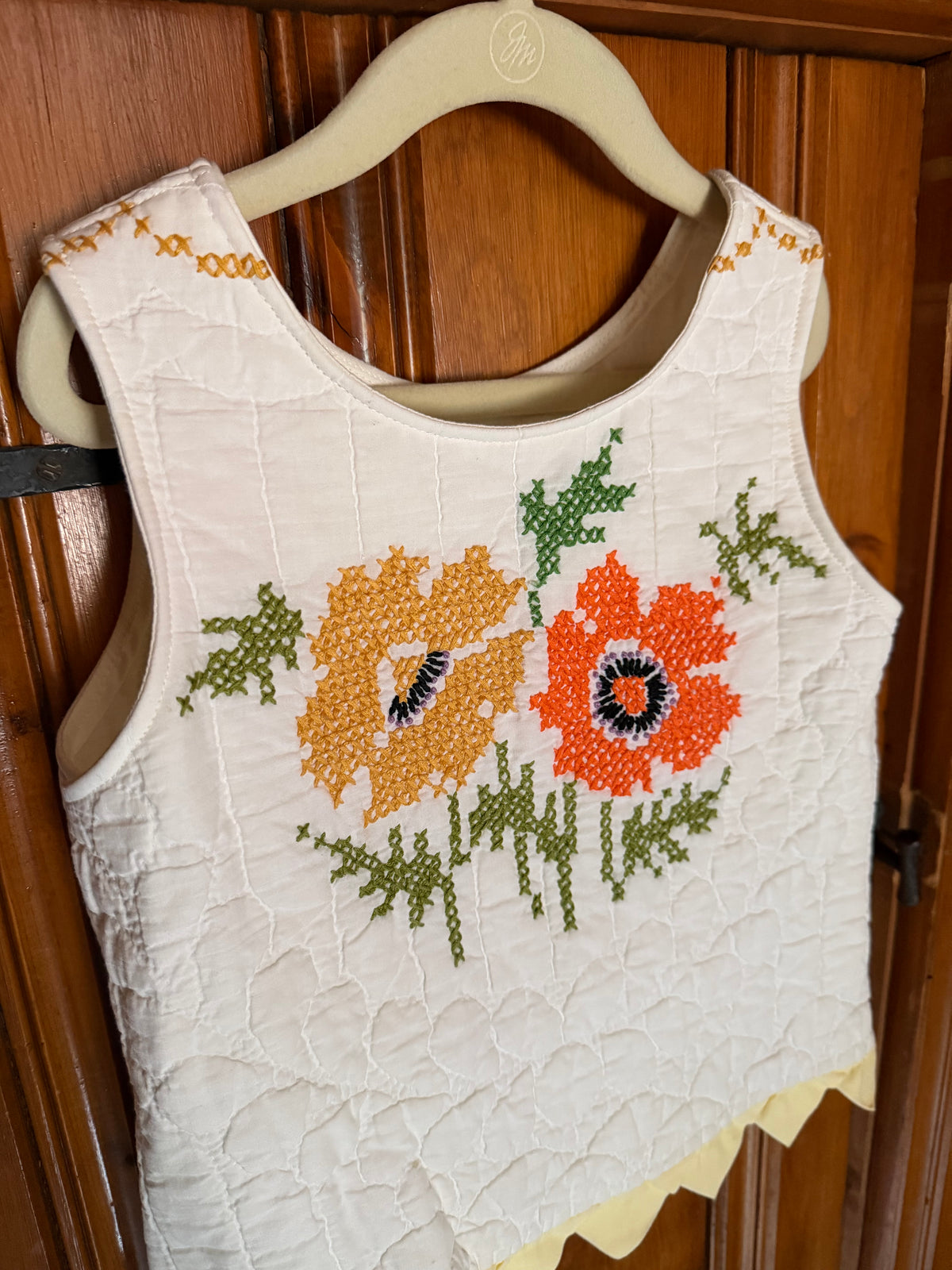 A sleeveless cream-colored quilted vest with embroidered orange and yellow flowers and green leaves, hanging on a wooden hanger against a dark wood paneled door.