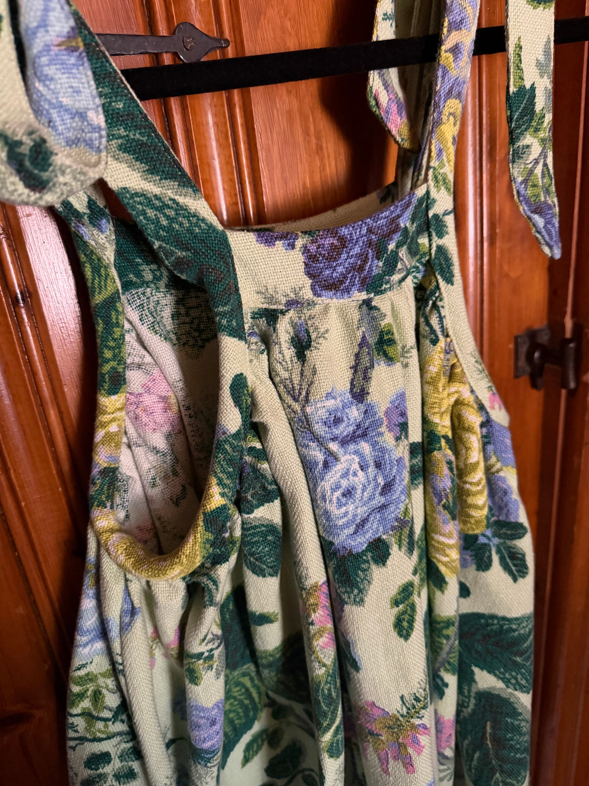 A close-up of a floral patterned dress or apron hanging on a black clothes hanger against a warm brown wooden door. The fabric features a detailed botanical print with purple, green, yellow, and pink flowers on a light beige background.