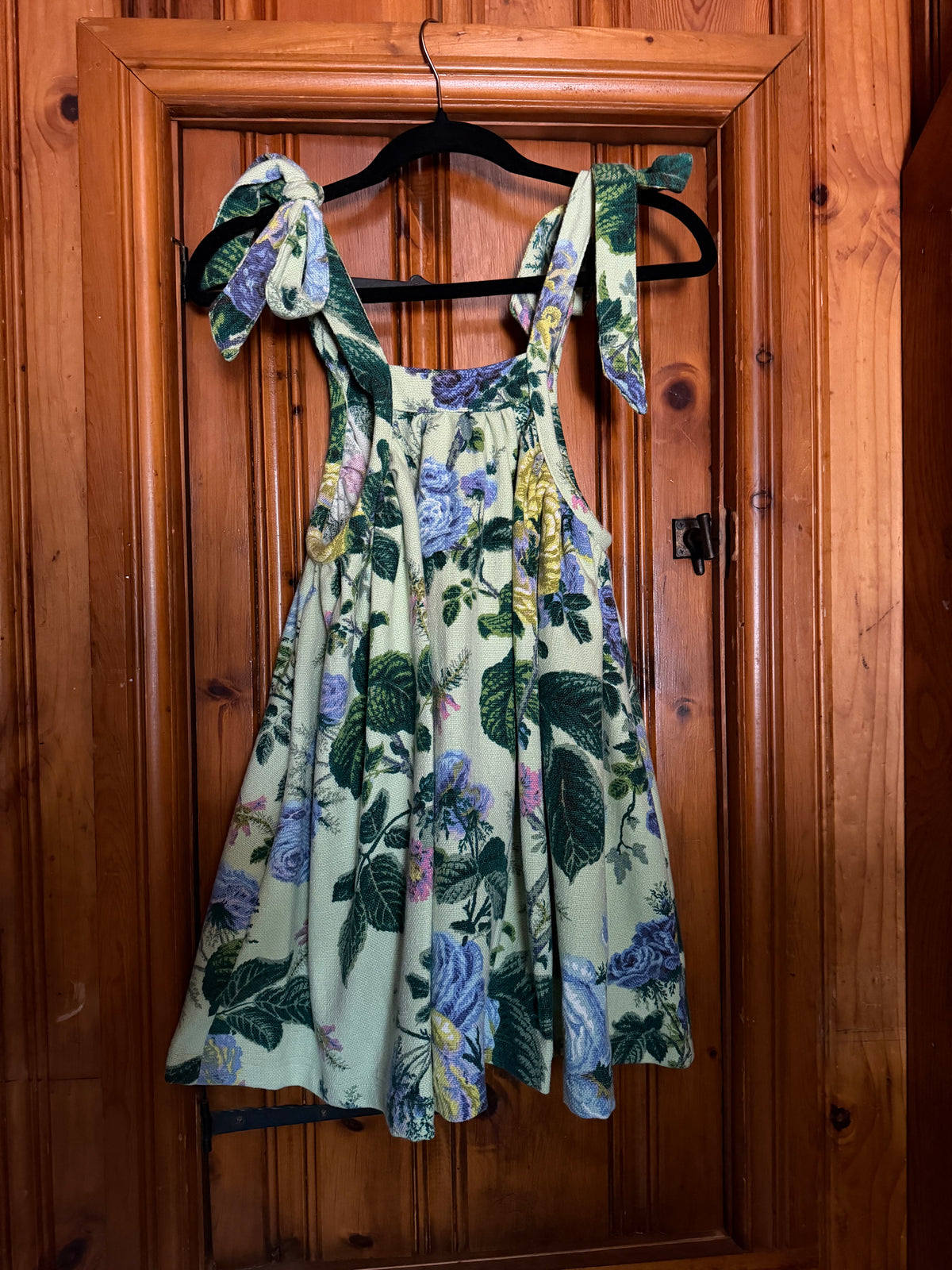 A sleeveless floral dress with tie-shoulder straps hangs on a black hanger against a rustic wooden door. The dress features a light sage green base with a vibrant print of blue-purple flowers, green leaves, and small yellow accents.