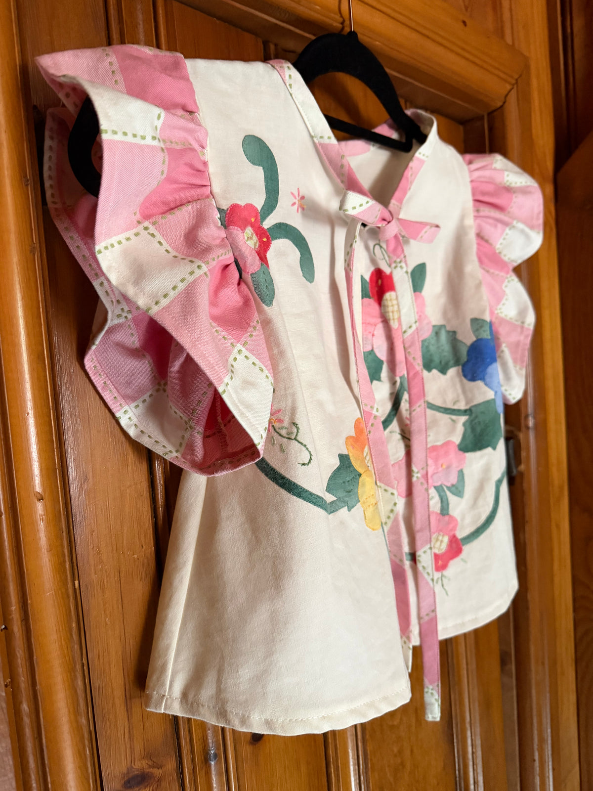 A pink and cream patchwork blouse with floral embroidery, ruffled sleeves, and a tie-neck, hanging on a black hanger against a wooden door.