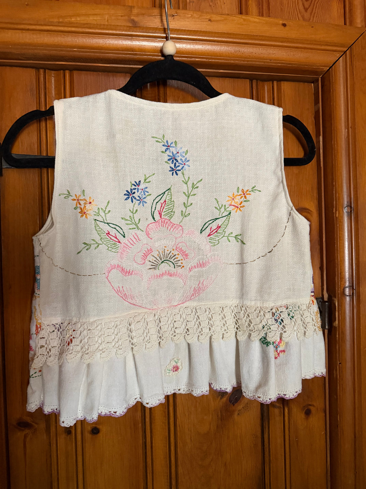 A sleeveless cream-colored linen vest with colorful floral embroidery and a lace hem, hanging on a black hanger against a wooden door.
