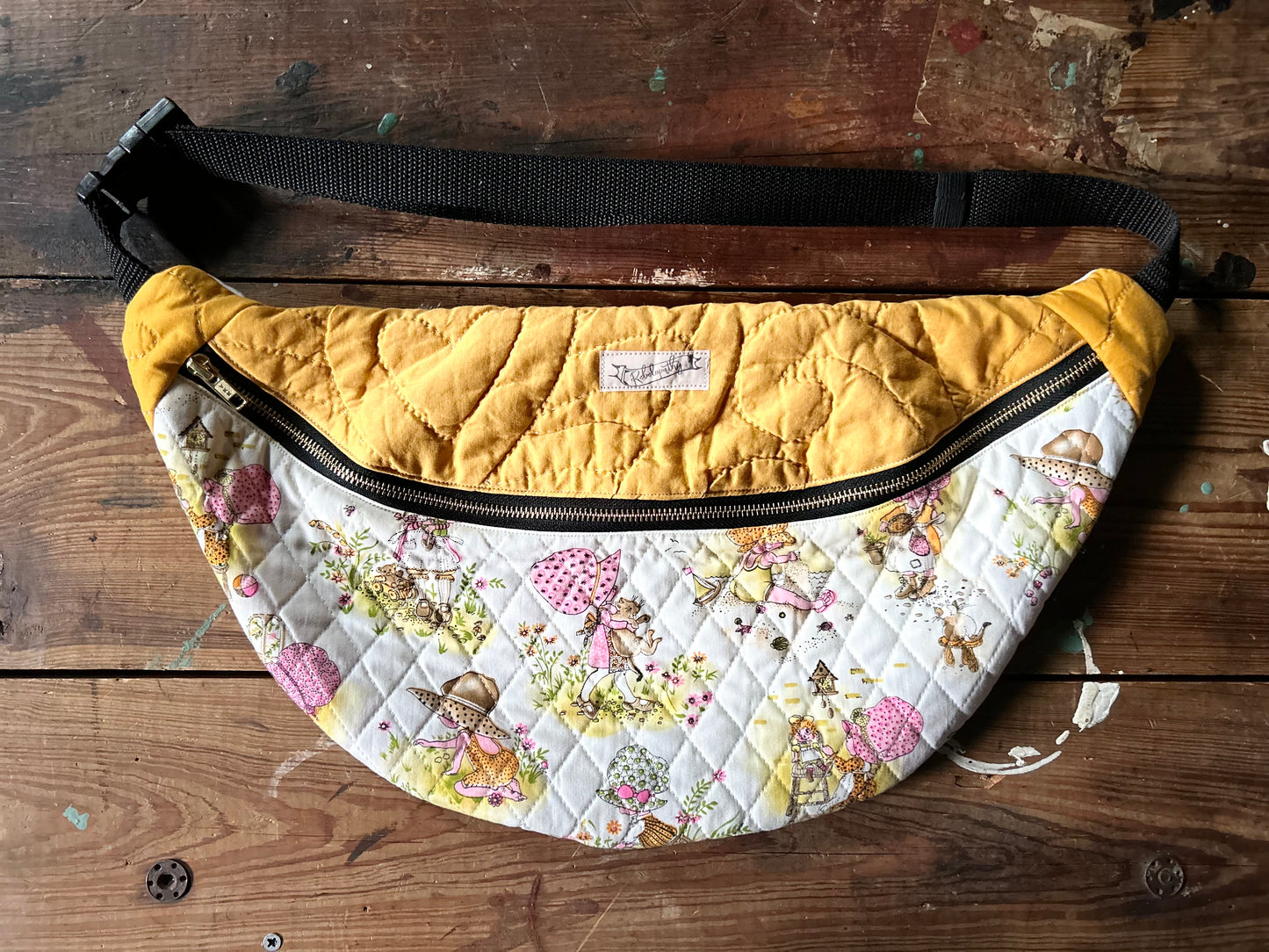 Multicolored Quilted Cross body bag in two tone vintage quilt and quilted fabric. Bottom plaid portion is predominantly white, yellow with touches of pink. The bottom quilt portion contains a vintage style girl in oversized hats in various activities such a carrying a large cat, gardening, playing in sand and playing with toys. Top portion is a vintage solid mustard yellow tone. Separated by a gold metal zipper and contains a buckle style closure with nylon strap. Photographed on a rustic wooden surface.