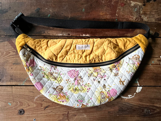 Multicolored Quilted Cross body bag in two tone vintage quilt and quilted fabric. Bottom plaid portion is predominantly white, yellow with touches of pink. The bottom quilt portion contains a vintage style girl in oversized hats in various activities such a carrying a large cat, gardening, playing in sand and playing with toys. Top portion is a vintage solid mustard yellow tone. Separated by a gold metal zipper and contains a buckle style closure with nylon strap. Photographed on a rustic wooden surface.