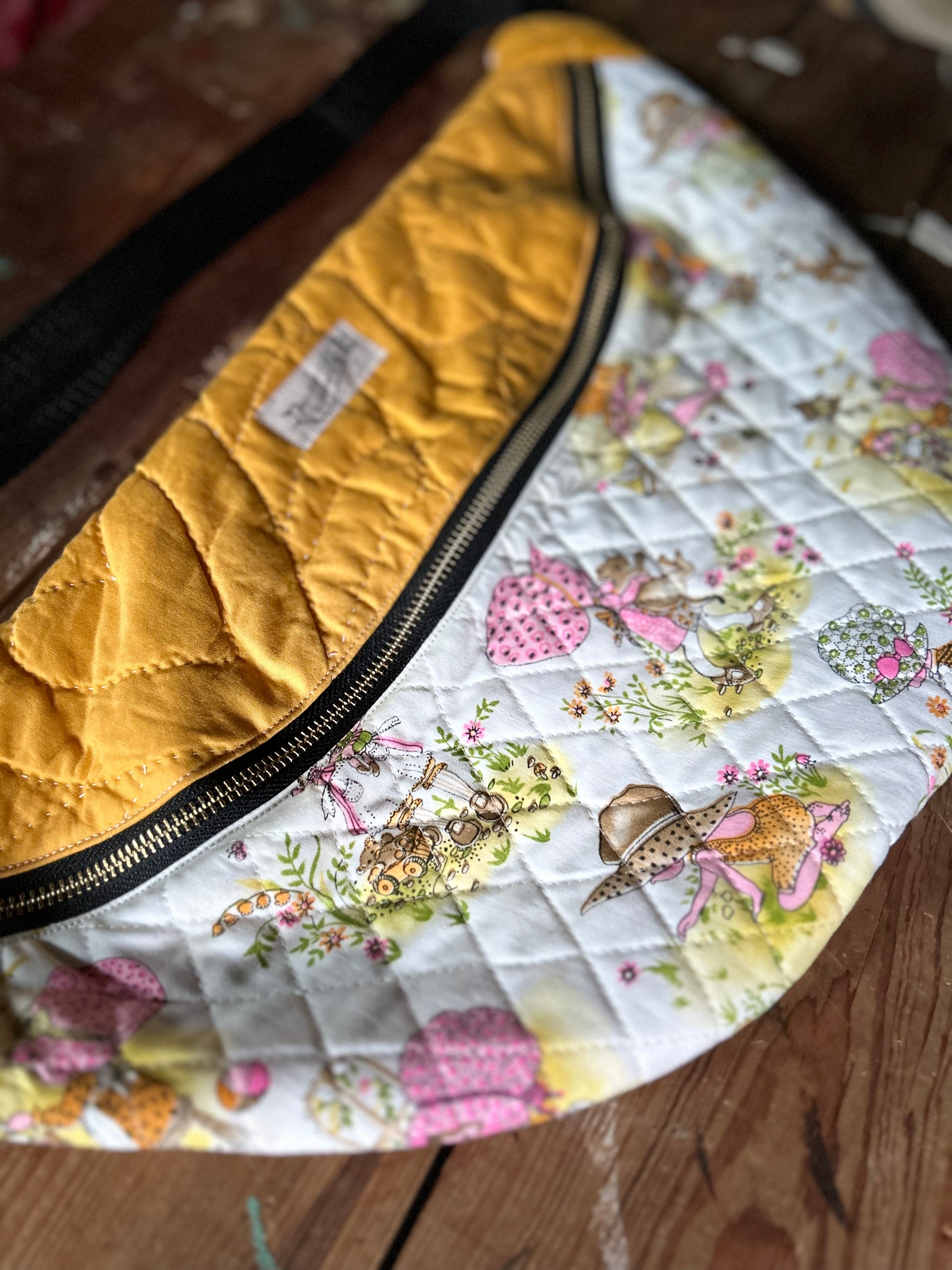 Closeup Quilted Cross body bag in two tone vintage quilt and quilted fabric. Bottom plaid portion is predominantly white, yellow with touches of pink. The bottom quilt portion contains a vintage style girl in oversized hats in various activities such a carrying a large cat, gardening, playing in sand and playing with toys. Top portion is a vintage solid mustard yellow tone. Separated by a gold metal zipper and contains a buckle style closure with nylon strap. Photographed on a rustic wooden surface.