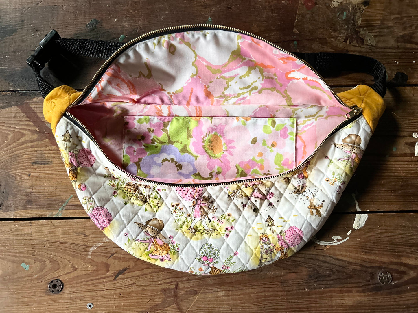 Inside view of Quilted Cross body bag in two tone vintage quilt and quilted fabric. Bottom plaid portion is predominantly white, yellow with touches of pink. The bottom quilt portion contains a vintage style girl in oversized hats in various activities such a carrying a large cat, gardening, playing in sand and playing with toys. Top portion is a vintage solid mustard yellow tone. Inside view is a vintage abstract pink floral print. Photographed on a rustic wooden surface.