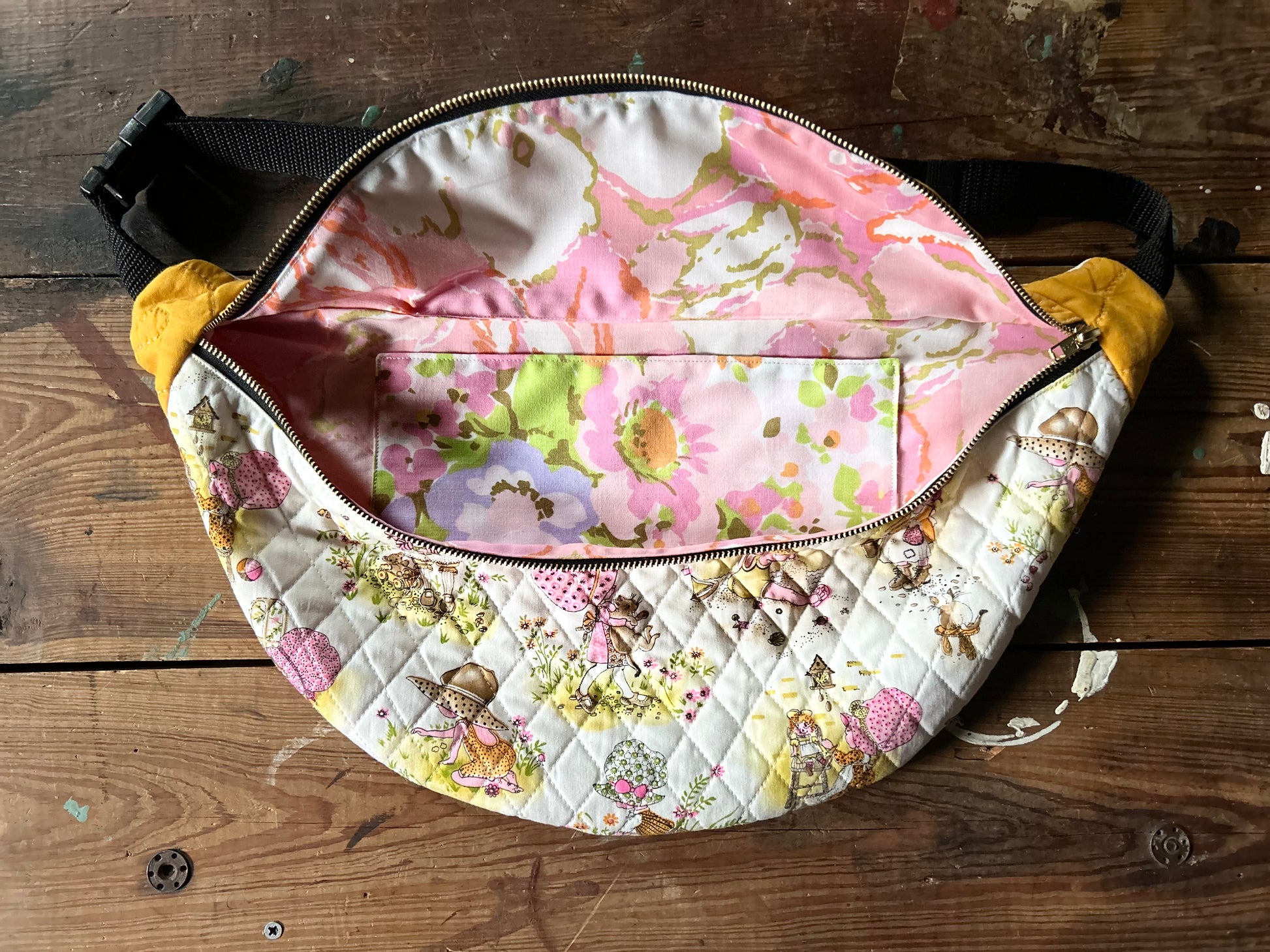 Inside view of Quilted Cross body bag in two tone vintage quilt and quilted fabric. Bottom plaid portion is predominantly white, yellow with touches of pink. The bottom quilt portion contains a vintage style girl in oversized hats in various activities such a carrying a large cat, gardening, playing in sand and playing with toys. Top portion is a vintage solid mustard yellow tone. Inside view is a vintage abstract pink floral print. Photographed on a rustic wooden surface.