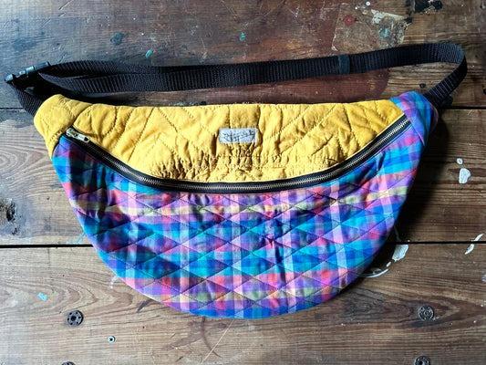 Multicolored Quilted Cross body bag in two tone vintage quilt and quilted fabric. Bottom plaid portion is predominantly blues, purples with touches pink, orange and yellow. Top portion is a vintage solid mustard yellow tone. Separated by a gold metal zipper and contains a buckle style closure with nylon strap. Photographed on a rustic wooden surface.