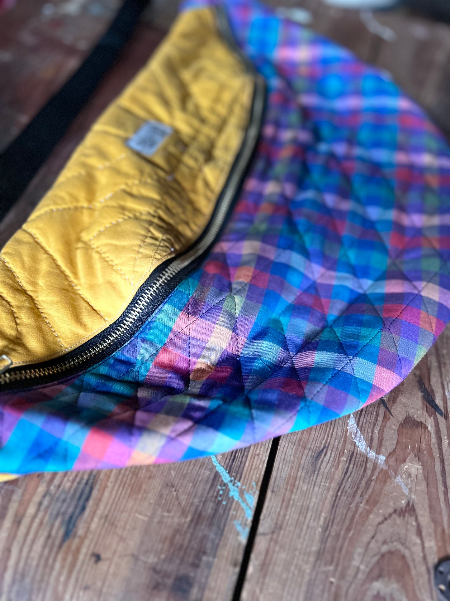 Close up of a multicolored Quilted Cross body bag in two tone vintage quilt and quilted fabric. Bottom plaid portion is predominantly blues, purples with touches pink, orange and yellow. Top portion is a vintage solid mustard yellow tone. Separated by a gold metal zipper and contains a buckle style closure with nylon strap. Photographed on a rustic wooden surface.