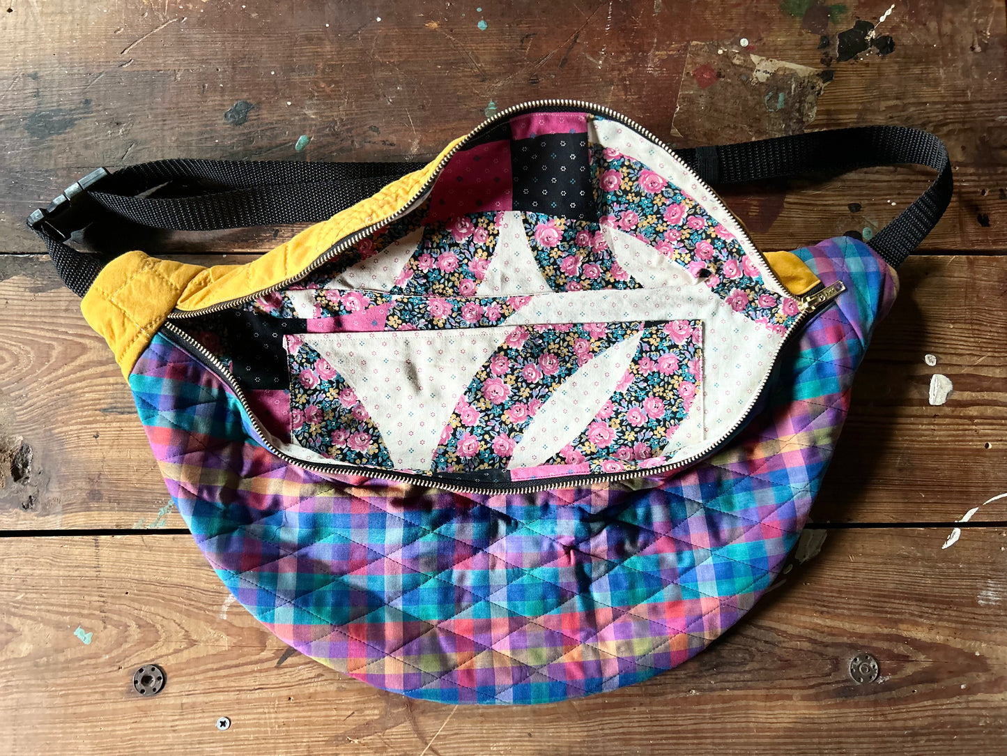 Multicolored Quilted Cross body bag unzipped in two tone vintage quilt and quilted fabric. Bottom plaid portion is predominantly blues, purples with touches pink, orange and yellow. Top portion is a vintage solid mustard yellow tone. Separated by a gold metal zipper and contains a buckle style closure with nylon strap. Inside contains a colorful faux wedding ring quilt design in pink, blue and yellow florals along with an oversized internal pocket. Photographed on a rustic wooden surface.