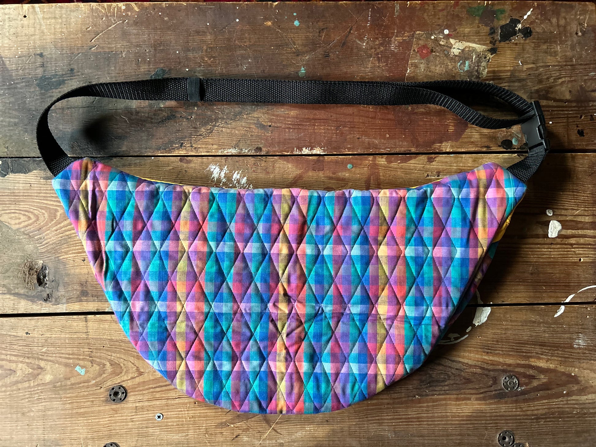Backside of a Multicolored Quilted Cross body bag in two tone vintage quilt and quilted fabric. Backside of bag is constructed only of the matching plaid fabric. Plaid portion is predominantly blues, purples with touches pink, orange and yellow. Contains a buckle style closure with nylon strap. Photographed on a rustic wooden surface.