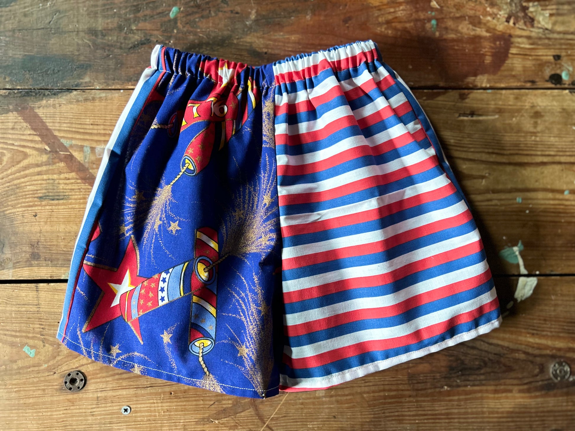 Pair of children’s red, white and blue shorts. Front panels are different with one side featuring lit firework fuses with gold details and 1776 firework. The other front panel features a red, white and blue stripe in a horizontal stripe.