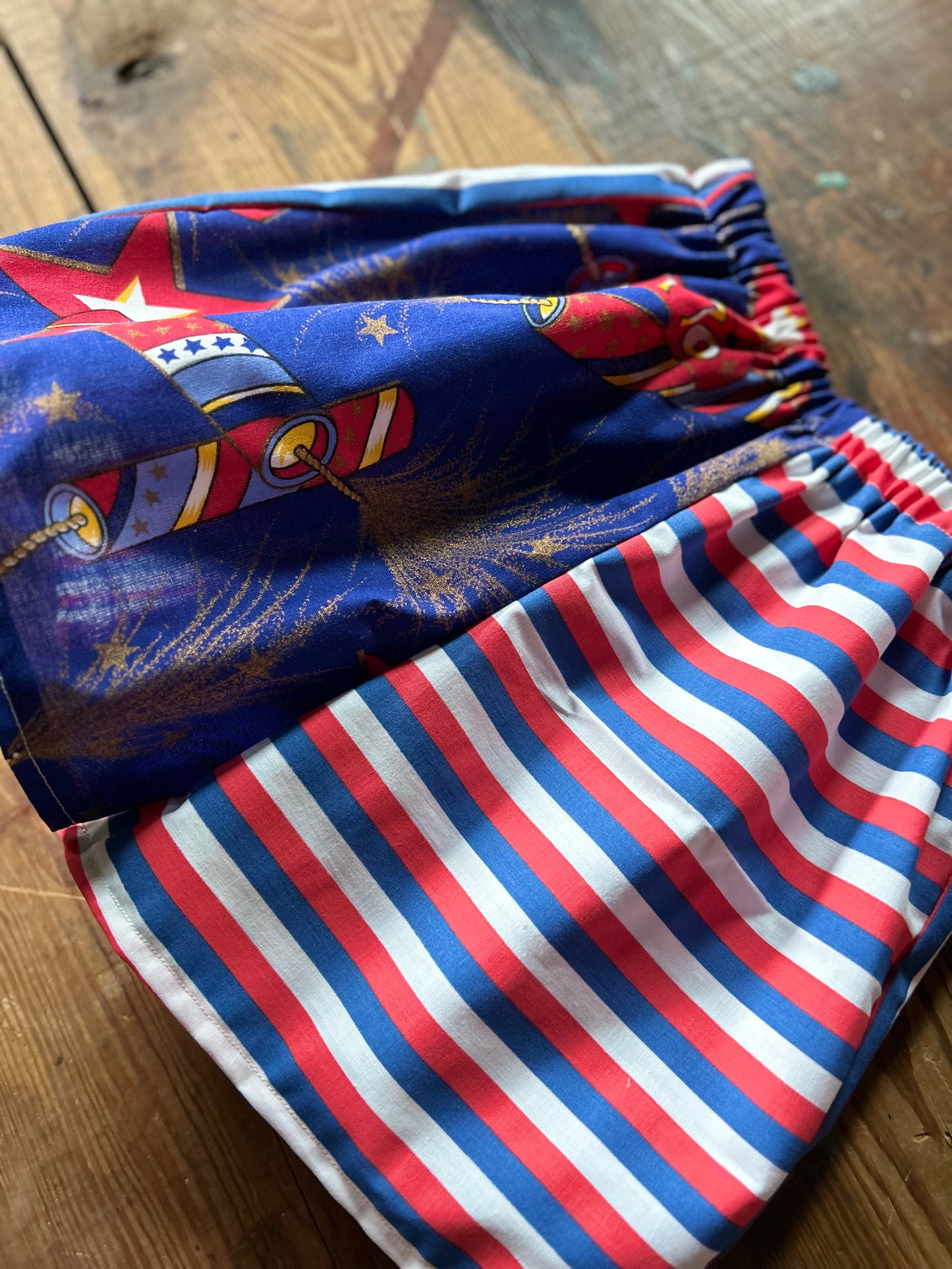 Closeup of Pair of children’s red, white and blue shorts to show sheerness in 1776 material. Front panels are different with one side featuring lit firework fuses with gold details and 1776 firework. The other front panel features a red, white and blue stripe in a horizontal stripe.