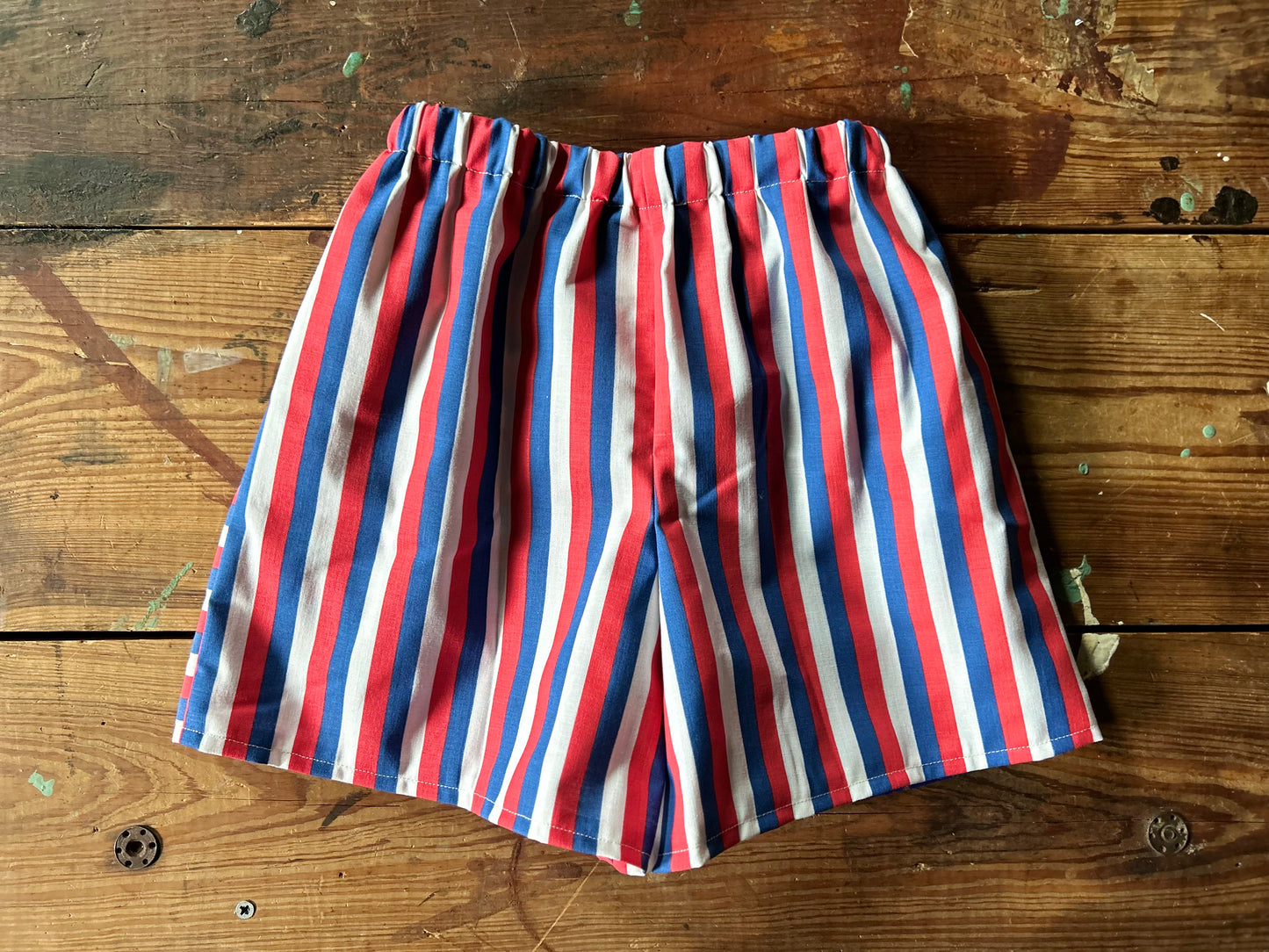 Back view of Pair of children’s red, white and blue shorts. Featuring a red, white and blue stripe in a vertical stripe.