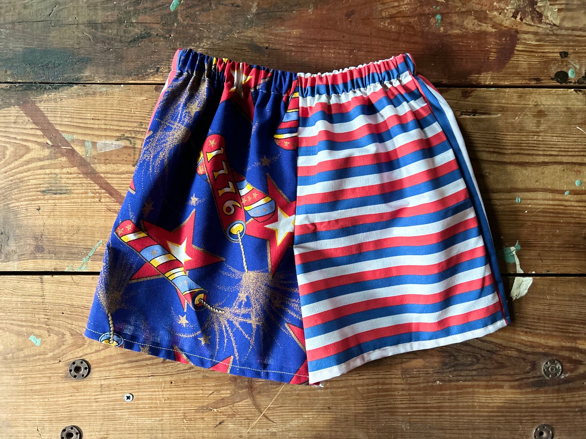 Pair of children’s red, white and blue shorts. Front panels are different with one side featuring lit firework fuses with gold details and 1776 firework. The other front panel features a red, white and blue stripe in a horizontal stripe.