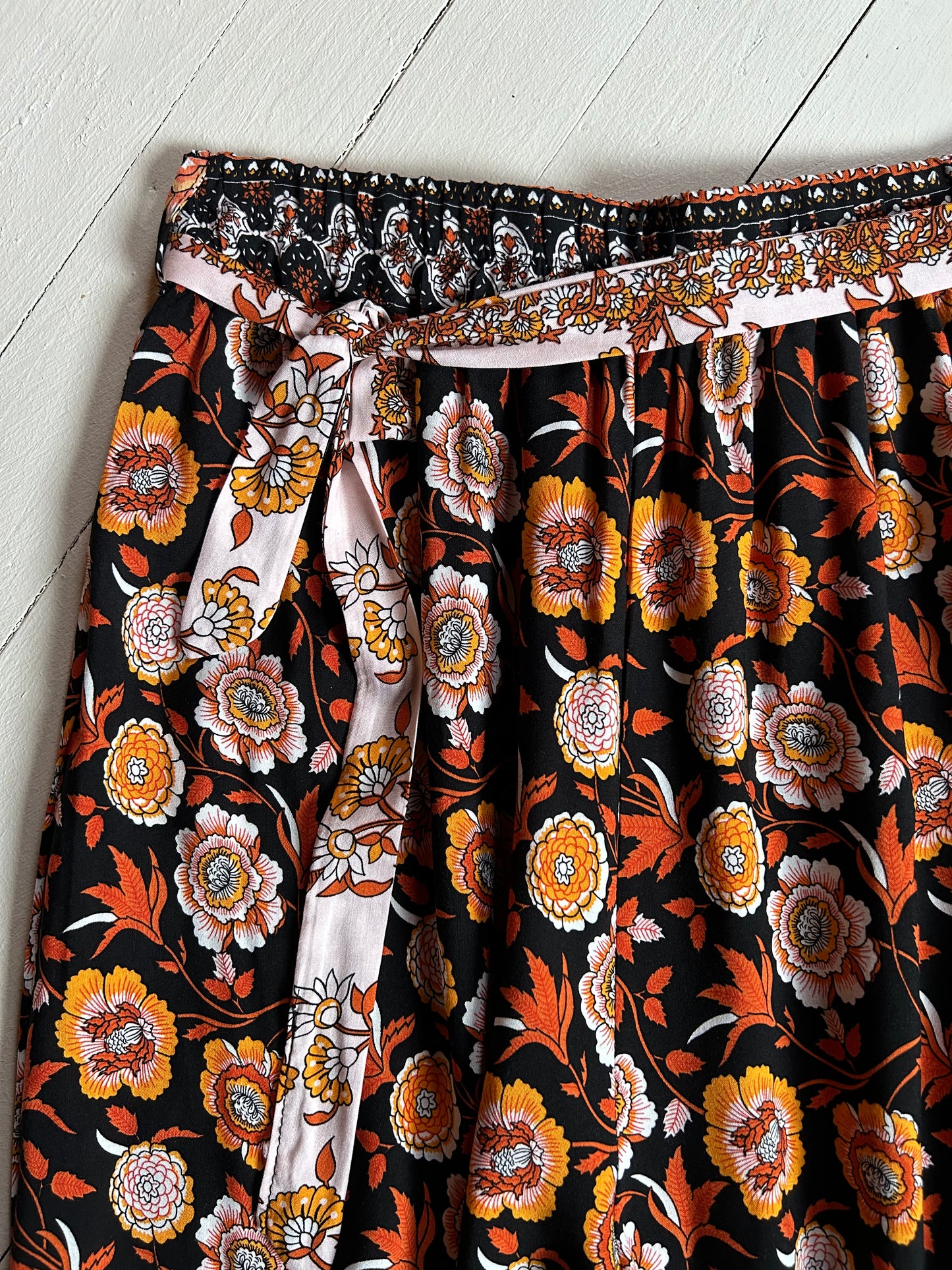 White wooded background. Closeup of front waist with tie of a Black elastic pants with orange and white floral. 