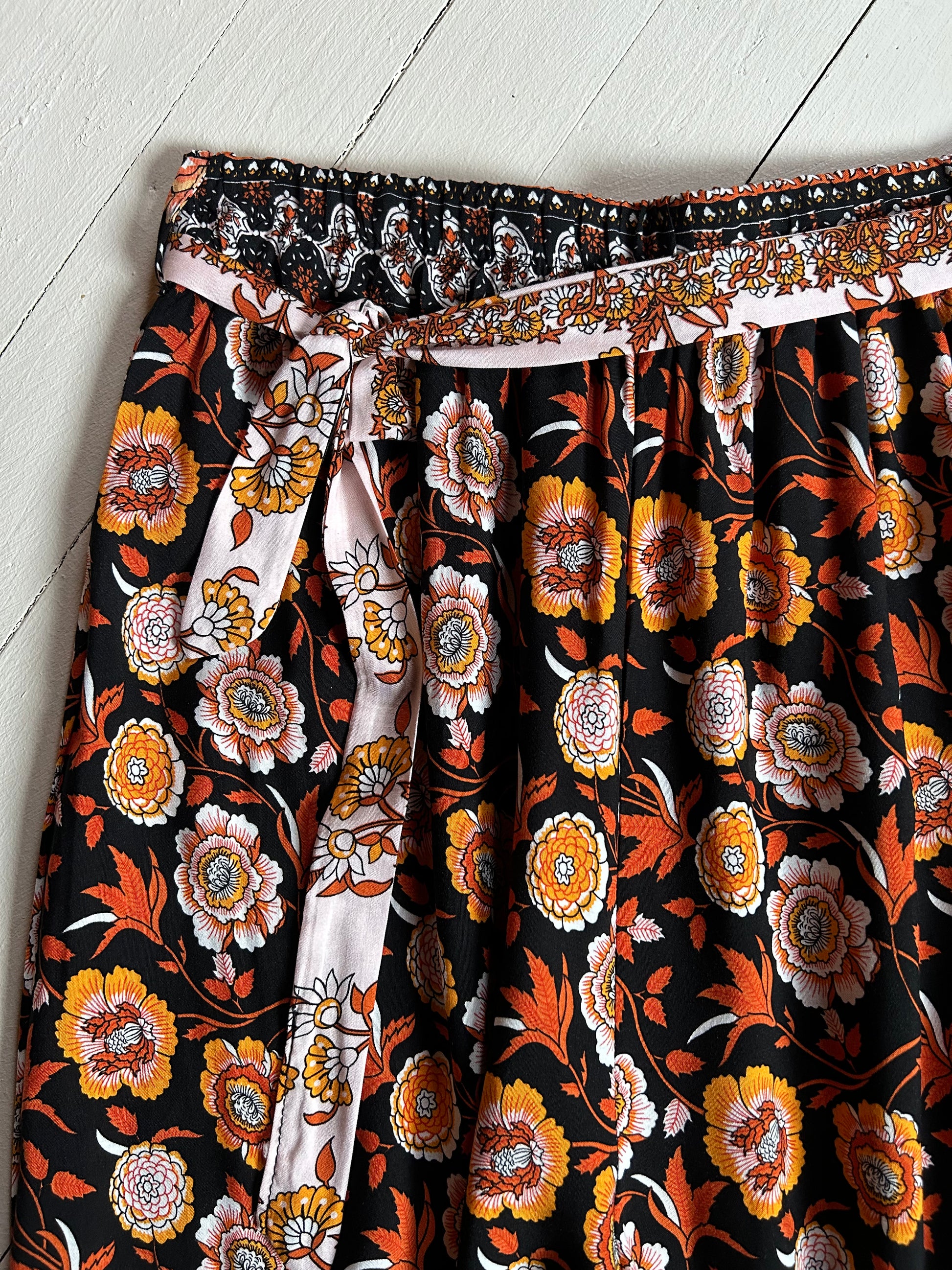 White wooded background. Closeup of front waist with tie of a Black elastic pants with orange and white floral. 